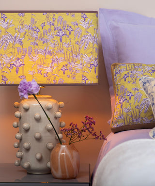 A styled bedroom scene featuring a table lamp with a yellow and lavender floral patterned shade and a textured beige ceramic base, alongside a matching decorative pillow on a bed with lavender bedding.