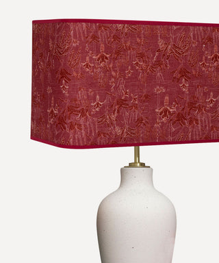 A table lamp with a textured, deep red rectangular shade featuring an embossed floral pattern and a gold-toned metal neck, mounted on a speckled white ceramic base against a light gray background.