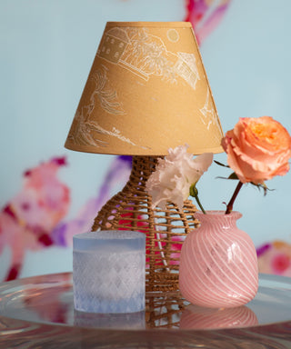 A styled tabletop scene with a tan patterned lampshade on a woven base, a pink ribbed glass vase, a pale pink flower, and a frosted candle against a soft pastel floral background.