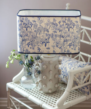 A table lamp with a cream fabric shade featuring a blue botanical toile pattern, set on a white rattan chair next to a matching pillow and a small glass vase of blue flowers.