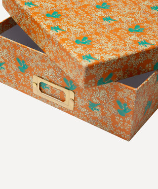 An orange storage box with a lid, featuring a white floral pattern and teal leaf motifs, with a gold metal handle, shown on a light gray background.
